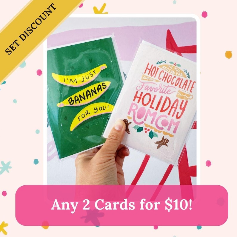 May include: Two greeting cards with a pink and white background. The card on the left has a green background with the text "I'm just bananas for you!" The card on the right has a white background with the text "It's the season for hot chocolate and your favorite holiday romcom." The text "Any 2 Cards for $10!" is displayed at the bottom of the image.