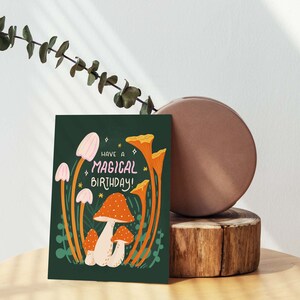 Greeting card with words "Have a magical birthday!" with trippy orange, beige, pink mushrooms (shiitake, enoki, toadstool, chanterelles) around with foliage and stars on a dark forest green background