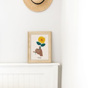 May include: A framed print of a yellow hibiscus flower being held by a brown hand making the shaka sign. The print has the word "Hawaii" written below the image.