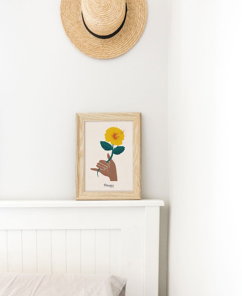 May include: A framed print of a yellow hibiscus flower being held by a brown hand making the shaka sign. The print has the word "Hawaii" written below the image.