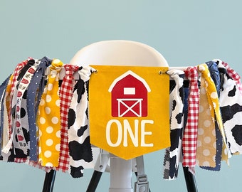 May include: A yellow and red barn-themed banner with the word "ONE" on it, hanging from a high chair. The banner is decorated with red and white gingham, yellow polka dots, and black and white cow print fabric.