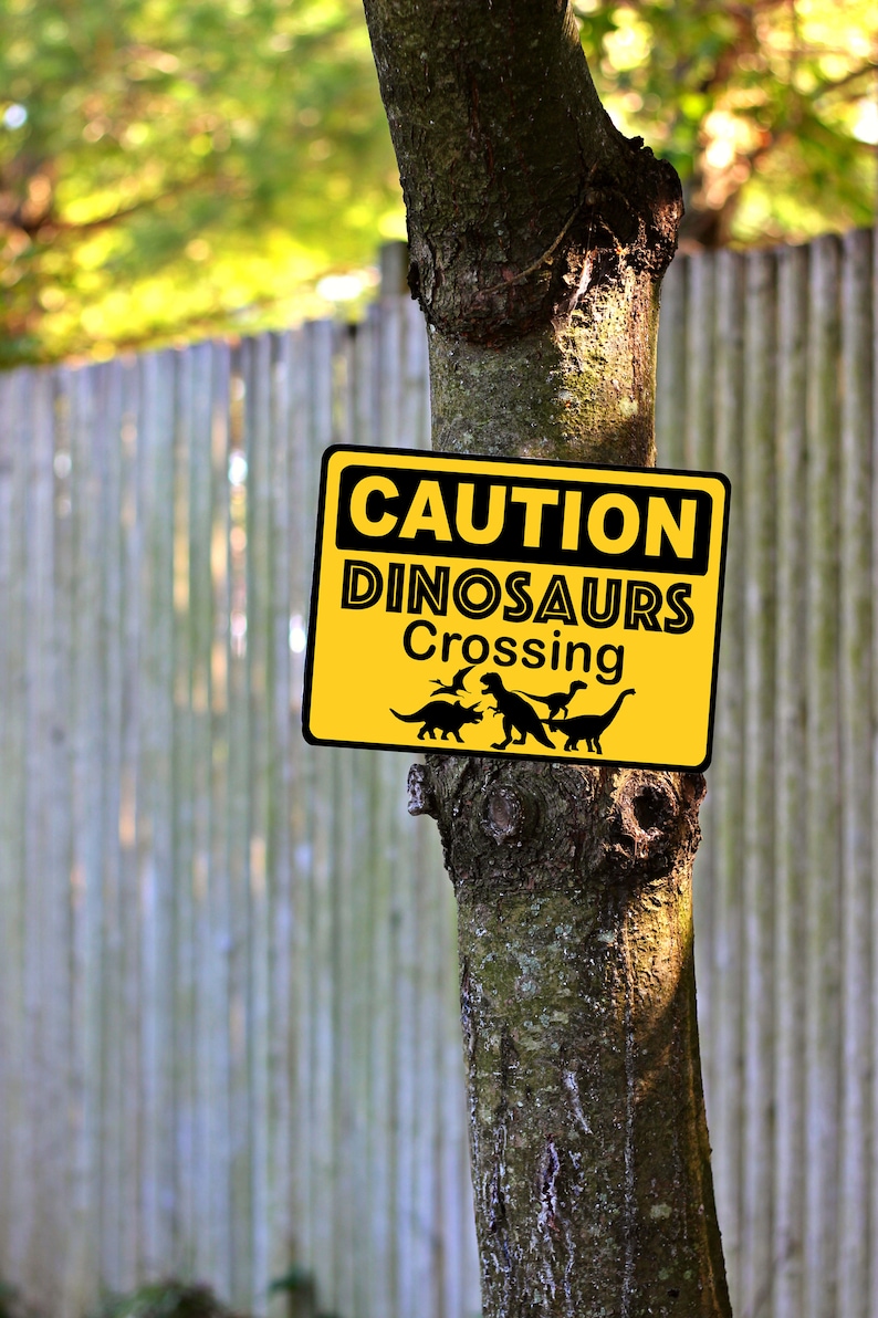 May include: A yellow caution sign with black text that reads "CAUTION DINOSAURS CROSSING". The sign is attached to a tree trunk with a wooden fence in the background.