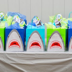 May include: A table with eight blue, green, and turquoise gift bags with shark faces on them. Each bag has a name tag with a different name: Sophia, Liam, Alexander, Shawn, Delilah, and three other names. The bags are filled with tissue paper.