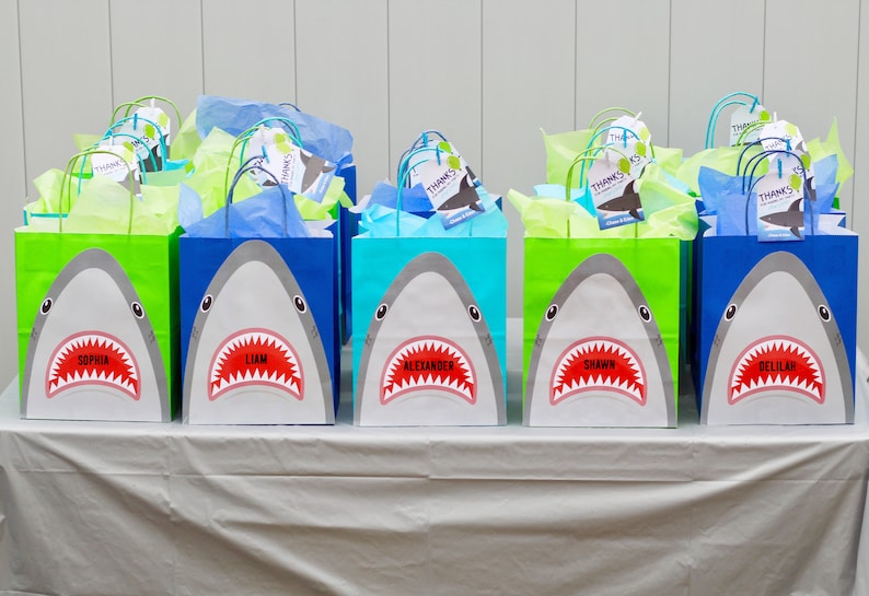 May include: A table with eight blue, green, and turquoise gift bags with shark faces on them. Each bag has a name tag with a different name: Sophia, Liam, Alexander, Shawn, Delilah, and three other names. The bags are filled with tissue paper.