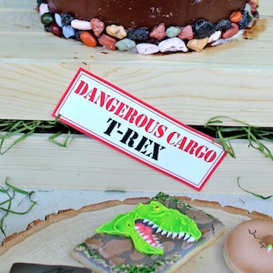 May include: A wooden crate with a sign that reads "Dangerous Cargo T-Rex" and green grass inside. The crate is sitting on a table with a dinosaur cookie and a brown cookie with a dinosaur footprint on it.