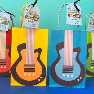 Puede incluir: Cuatro bolsas de regalo coloridas con diseños de guitarra y etiquetas "Gracias por hacer de mi fiesta un rock! -Daniel". Las bolsas son roja, amarilla, azul y verde.