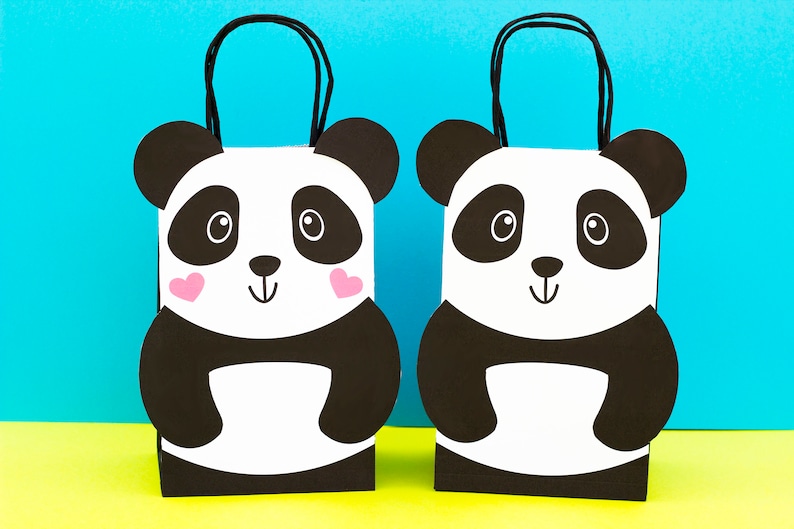 May include: Two black and white panda bear gift bags with pink hearts on their cheeks. The bags have black handles and are standing on a yellow surface.