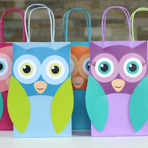Op de afbeelding: Vijf kleurrijke papieren cadeauzakjes met uilontwerpen. De zakjes zijn roze, blauw, oranje, paars en groen. Elk zakje heeft een wit uilgezicht met zwarte ogen en een gele snavel. De uilen hebben groene vleugels.