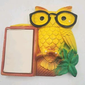 May include: A yellow owl-shaped wall decoration wearing black glasses, holding a notepad. The owl has textured feathers and is perched on a branch with green leaves. The notepad has a blank, white surface.