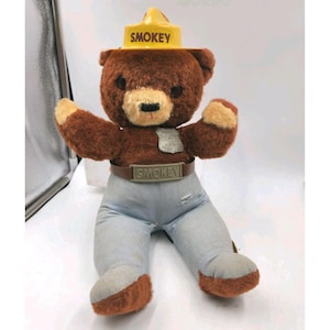 May include: A plush Smokey Bear doll with a brown body, tan snout, and blue denim pants. The doll wears a yellow hat and a brown belt, both with the word "SMOKEY". A badge is attached to the chest.