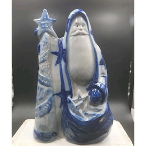 May include: A ceramic Santa Claus figurine in white and blue. The figure is holding a staff with a star on top and a sack. The Santa Claus is wearing a hooded robe with blue star patterns.