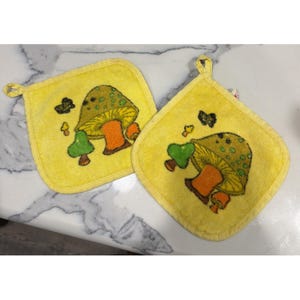 May include: Two square yellow potholders featuring a repeating graphic print of a large olive green mushroom with an orange stem, a smaller bright green mushroom, and a tiny orange mushroom, each with a small fabric loop for hanging.