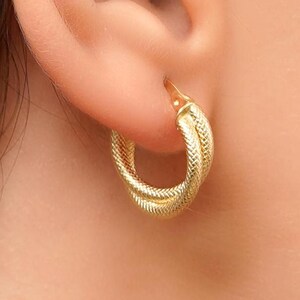 Może przedstawiać: Złote kolczyki koła z  plecionym wzorem. Kolczyki są noszone w uchu osoby.