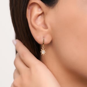 May include: Gold hoop earrings with a small sun charm featuring a clear crystal in the center.