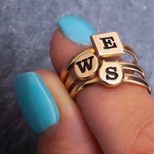 Puede incluir: Anillos de oro con dijes de letras negras que deletrean "WSE". Los anillos est&aacute;n apilados en un dedo con esmalte de u&ntilde;as azul claro.