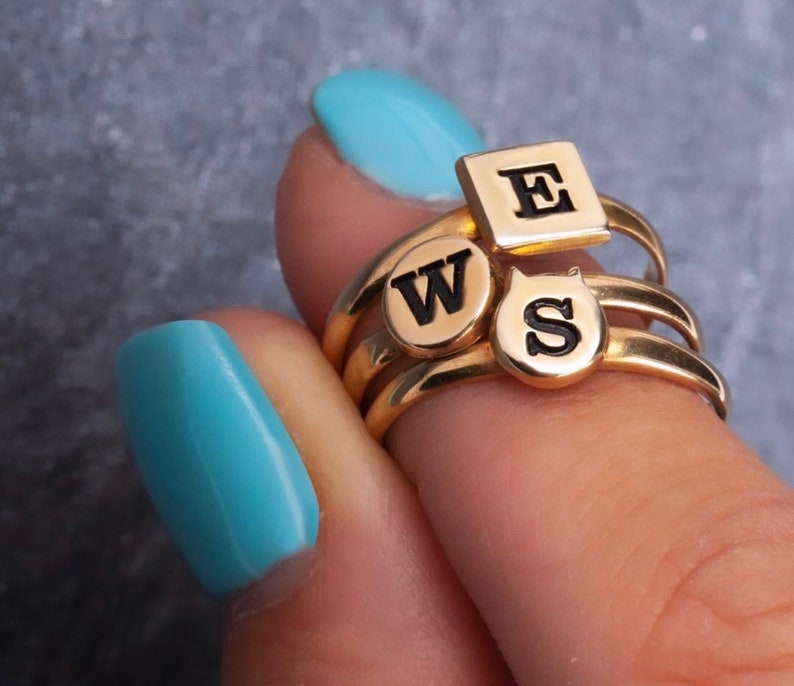 Puede incluir: Anillos de oro con dijes de letras negras que deletrean "WSE". Los anillos est&aacute;n apilados en un dedo con esmalte de u&ntilde;as azul claro.