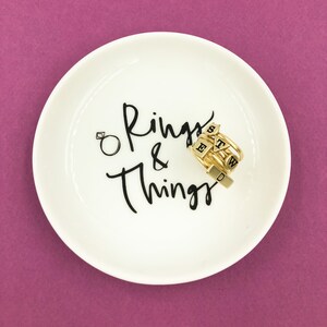 Puede incluir: Plato de cer&aacute;mica blanco para anillos con la inscripci&oacute;n "Rings & Things" en negro. Un conjunto de anillos de oro con abalorios de letras negras est&aacute; en el plato.