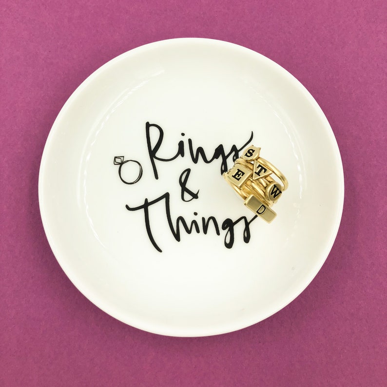 Puede incluir: Plato de cer&aacute;mica blanco para anillos con la inscripci&oacute;n "Rings & Things" en negro. Un conjunto de anillos de oro con abalorios de letras negras est&aacute; en el plato.