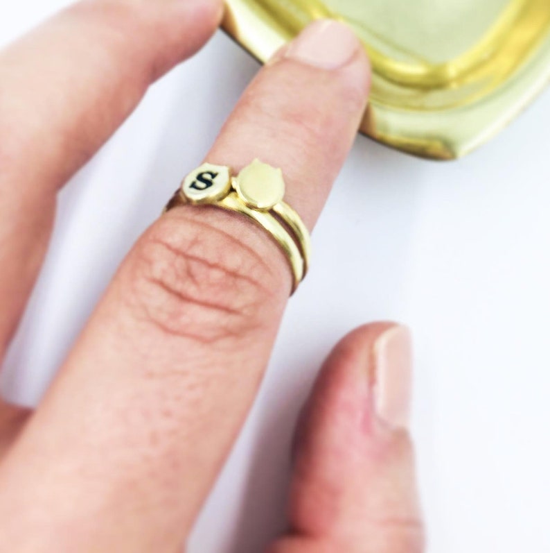 Puede incluir: Dos anillos de oro en un dedo. Un anillo tiene un peque&ntilde;o encanto redondo de oro con una letra "S" negra en &eacute;l. El otro anillo tiene un peque&ntilde;o encanto redondo de oro con forma de cabeza de gato.