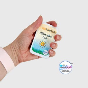 May include: A white rectangular tin of "Affirmation Cards" is held in a hand. The tin has a colourful design with a sun and grass. The text "Pocket Pal Tin" and "Affirmation Cards" are visible. The Planit Visuals logo is in the lower right.