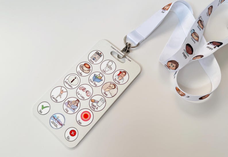 May include: White lanyard with colorful cartoon faces printed on it. A white plastic card with a variety of colorful icons and text is attached to the lanyard. The text on the card includes "Food/Drink", "Toilet", "Information", "Ear Loops", "Sleep", "Pain", "Yes", "No", "Hug", "Help", "Drink", "Eat", and "Coffee".