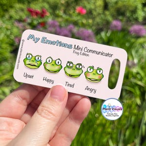 May include: A light pink Mini Communicator card with the text "My Emotions Frog Edition." The card features four green frog illustrations representing emotions: upset, happy, tired, and angry. The card is held in a hand.