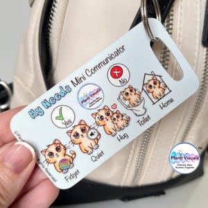 May include: A white and blue visual communication card with a cat theme. The card has images and text for common needs such as "Yes", "No", "Fidget", "Quiet", "Hug", "Toilet", and "Home". The card is attached to a white bag with a zipper.