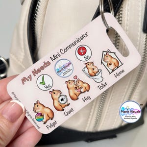 May include: A white and pink Mini Communicator tag with a silver keyring. The tag features cartoon hamsters and the words "My Needs Mini Communicator". Icons include "Yes", "No", "Fidget", "Quiet", "Hug", "Toilet", and "Home".
