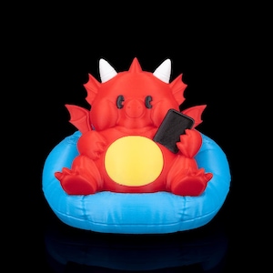 May include: A red dragon figurine with white horns and small wings, holding a black rectangle, possibly a phone. The dragon is seated on a blue cushion with a yellow circle on its belly. The background is black.
