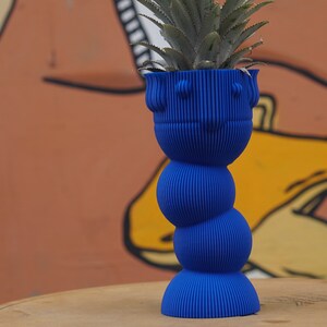 Puede incluir: Un macetero de cerámica azul con una superficie texturizada y un diseño redondeado y segmentado. Una planta de piña está creciendo en el macetero.