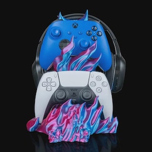 May include: A blue Xbox controller with devil horn accents sits above a white PlayStation controller. A black headset is behind the controllers. A decorative base with a flame design in pink, blue, and purple supports the controllers.