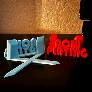 Puede incluir: Un letrero impreso en 3D que dice "Now Playing" en rojo y "Home" en azul. El letrero está hecho de plástico y tiene un diseño moderno.