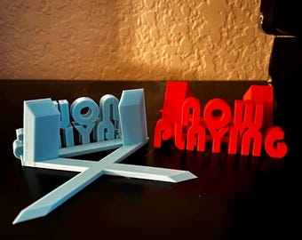May include: A 3D printed sign that says "Now Playing" in red and "Home" in blue. The sign is made of plastic and has a modern design.