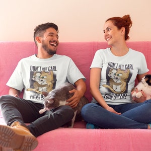Könnte beinhalten: Zwei Personen sitzen auf einem rosa Sofa und tragen weiße T-Shirts mit einem Grafikdruck von zwei Katzen und dem Text "Lass Carl nicht dein 9-to-5-Leben ruinieren".