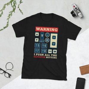 Op de afbeelding: Zwart T-shirt met een witte afbeelding van een liftknop paneel. De afbeelding zegt "WAARSCHUWING" en bevat een verscheidenheid aan knoppen, waaronder pijlen omhoog en omlaag, een bel en de nummers 5 tot en met 8. De tekst onder de afbeelding zegt "IK DRUK OP ALLE LIFTKNOPPEN".