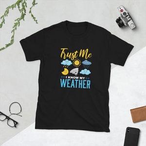 Puede incluir: Camiseta negra con un gráfico blanco que dice "Trust Me I Know My Weather". El gráfico incluye un sol, nubes, rayos y un tornado.
