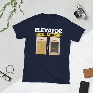 May include: Navy blue t-shirt with the text "Elevator Whisperer" in white and yellow. Two cartoon images of lifts are below the text.