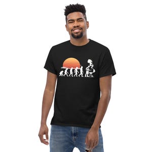 May include: Black t-shirt featuring a white graphic of human evolution leading to a chef grilling, with a sunset in the background. The shirt is made of a soft material and is suitable for casual wear.
