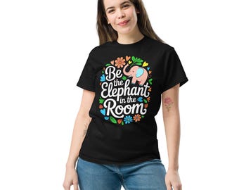 May include: Black t-shirt with the phrase "Be the Elephant in the Room" in white script, with a pink elephant graphic and colorful floral accents. The shirt is made of a soft material and is being worn by a person.