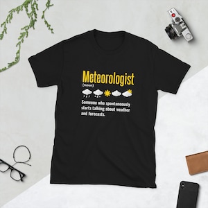 Può includere: Maglietta nera con una definizione gialla della parola "Meteorologo" con icone meteorologiche. La definizione recita: "Qualcuno che inizia spontaneamente a parlare del tempo e delle previsioni."