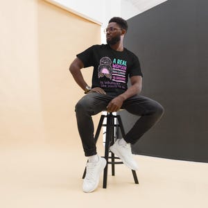 May include: A black t-shirt with the text "A Real Woman Has Curves Is Skinny Is Whatever She Wants To Be" in pink and blue. The shirt features a graphic of a woman with a pink heart. The person is wearing black jeans and white trainers.