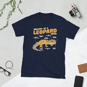 Może przedstawiać: Koszulka w kolorze granatowym z grafiką przedstawiającą gekona lamparciego i schematem jego anatomii. Tekst "Anatomy of a Leopard Gecko" jest nadrukowany na żółto w górnej części grafiki. Etykiety schematu obejmują "Mighty Suction Cups", "Shimmering Armor", "The Mastermind", "Gossip Antenna", "Soulful Peepers", "Cutie Ejector", "Bottomless Pit" i "Sticky Sader".