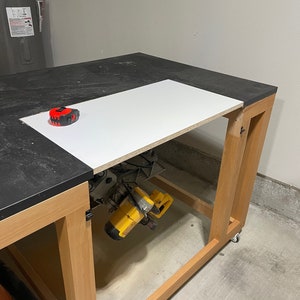 Pu&ograve; includere: Un banco da lavoro in legno con un piano nero e uno scaffale bianco. Uno strumento elettrico giallo &egrave; appeso sotto lo scaffale. Il banco da lavoro &egrave; su ruote e ha un metro a nastro sulla parte superiore.