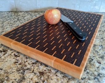 May include: A wooden cutting board with a walnut and maple inlay design. The board has a light brown border and a dark brown center with a woven pattern. A knife and an apple are on the board.