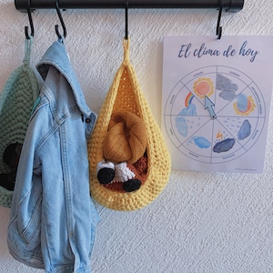 Può includere: Un cesto di stoccaggio appeso in crochet giallo con un peluche marrone e bianco al suo interno. Il cesto è appeso a un gancio di metallo nero. Una giacca di jeans blu e un cesto di crochet verde sono appesi a ganci accanto. Un foglio di carta bianco con il testo "El clima de hoy" e una ruota del tempo è sul muro dietro i cesti.