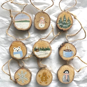May include: Nine round wooden slice ornaments with jute twine hangers. Hand-painted winter scenes include a landscape, snowman, pine trees, polar bear, snowy cabin, mug of cocoa, snowflake, gingerbread person, and a penguin wearing a pink scarf.