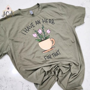May include: Olive green t-shirt with the phrase "I HAVE AN HERB FOR THAT" in black lettering. The shirt features a whimsical illustration of pink flowers in a teacup.