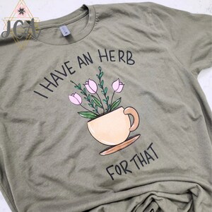 May include: Olive green t-shirt with the phrase "I HAVE AN HERB FOR THAT" in black lettering. A teacup illustration contains pink flowers and green leaves. The shirt is made of soft material.
