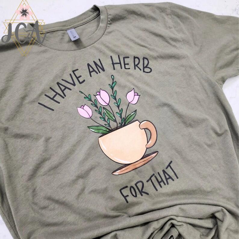 May include: Olive green t-shirt with the phrase "I HAVE AN HERB FOR THAT" in black lettering. A teacup illustration contains pink flowers and green leaves. The shirt is made of soft material.