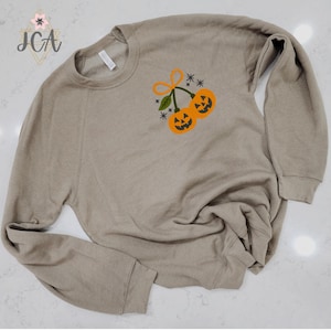 May include: A tan-colored sweatshirt with a crew neck and long sleeves. The front features an embroidered design of two orange jack-o'-lanterns with green leaves and an orange bow. The design is accented with small blue stars. The sweatshirt is made of a soft material.